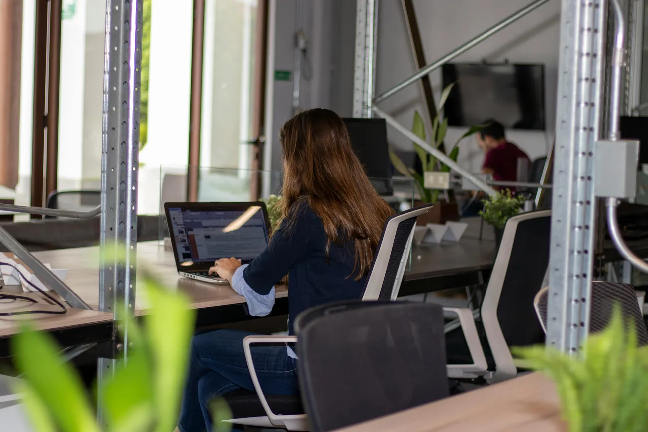 A woman with long hair is working on a laptop in a modern office with metal beams and plants. The atmosphere is focused and professional.
