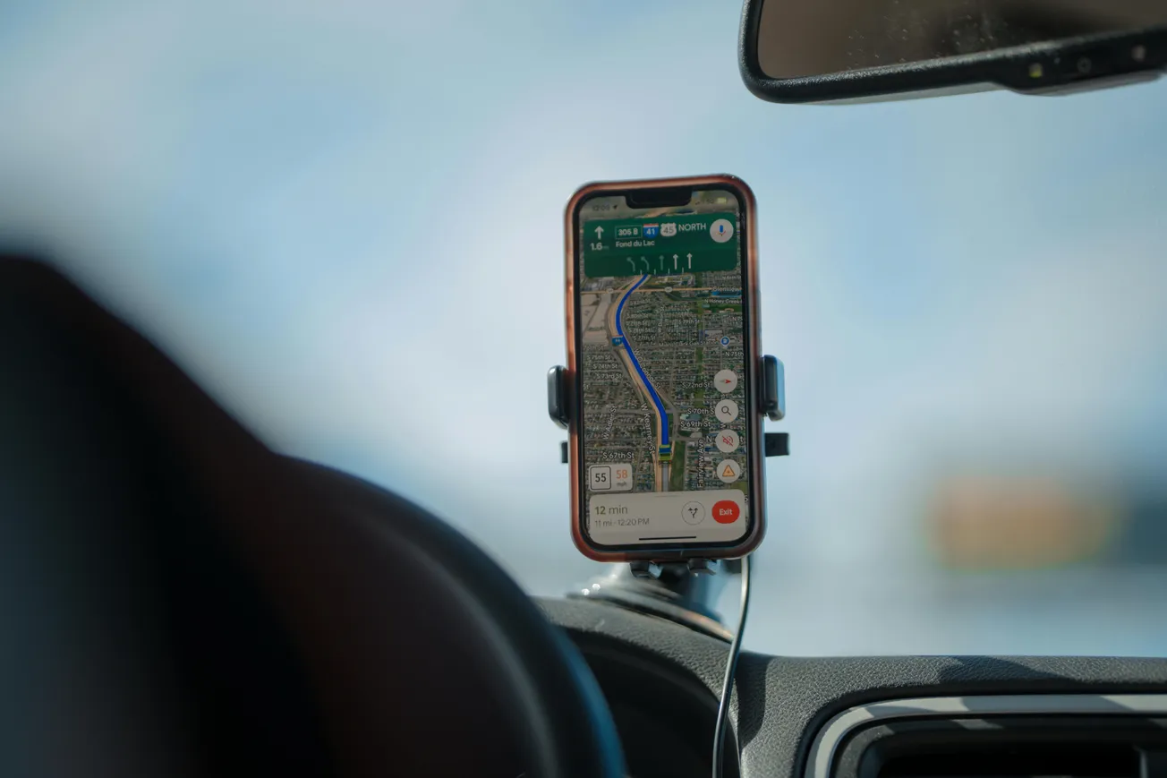 Smartphone with GPS map app in car mount shows navigation on a sunny day. Blue route, clear sky visible through windshield, suggesting travel.