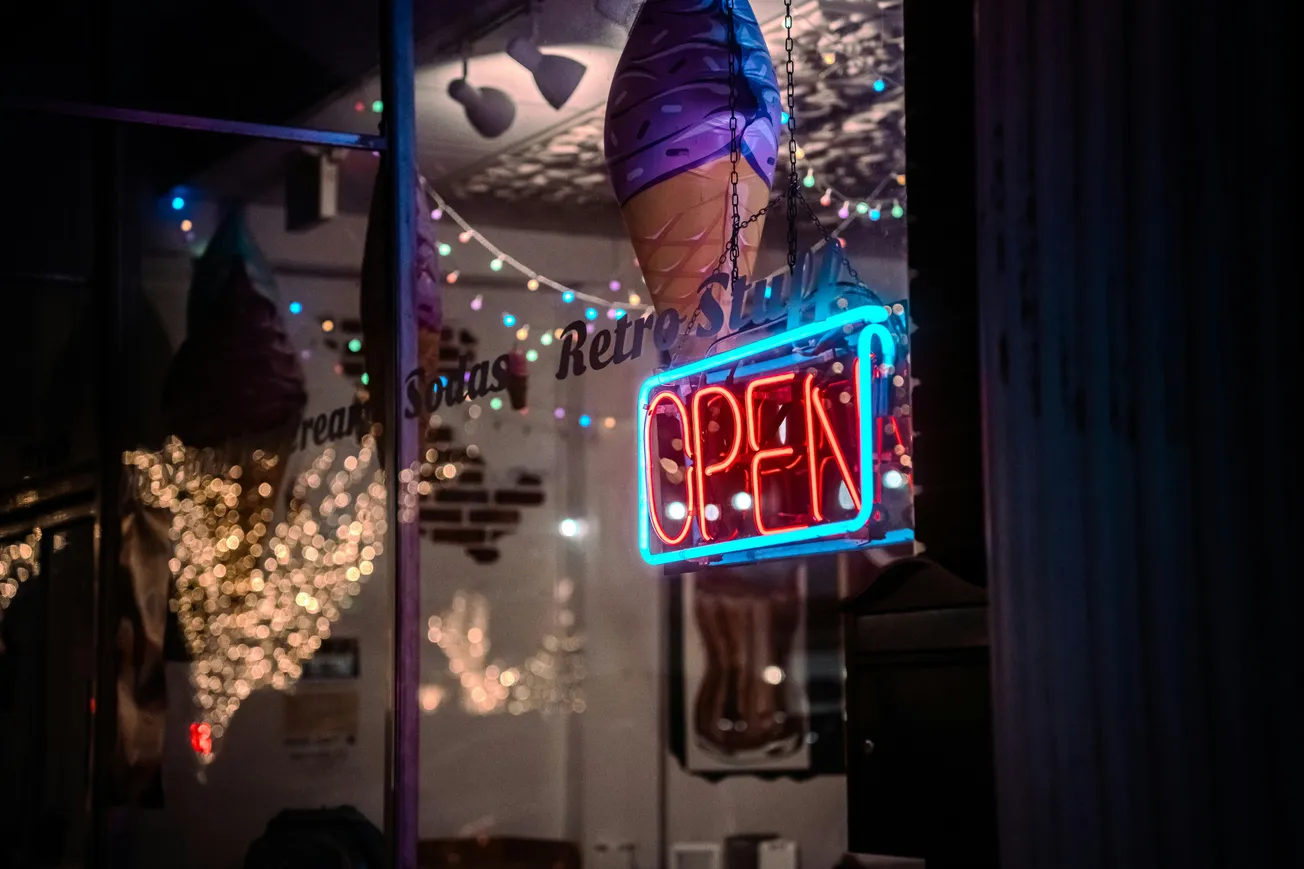 A neon "OPEN" sign glows in red and blue outside a dimly lit storefront, with a large ice cream cone decoration and colorful string lights inside.