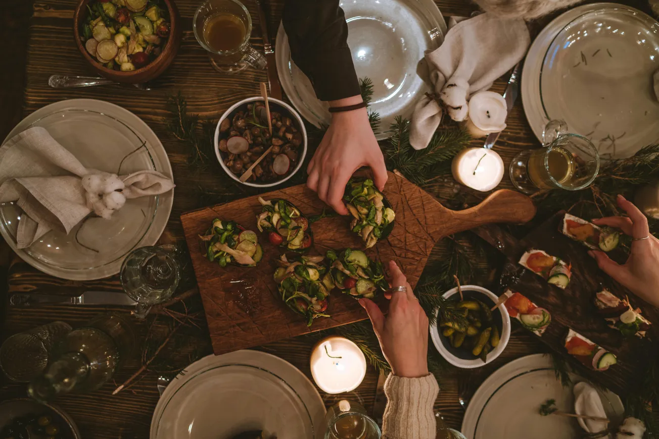 A cozy dinner setting with hands serving bruschetta from a wooden board, surrounded by lit candles, empty plates, and green decor on a rustic table.