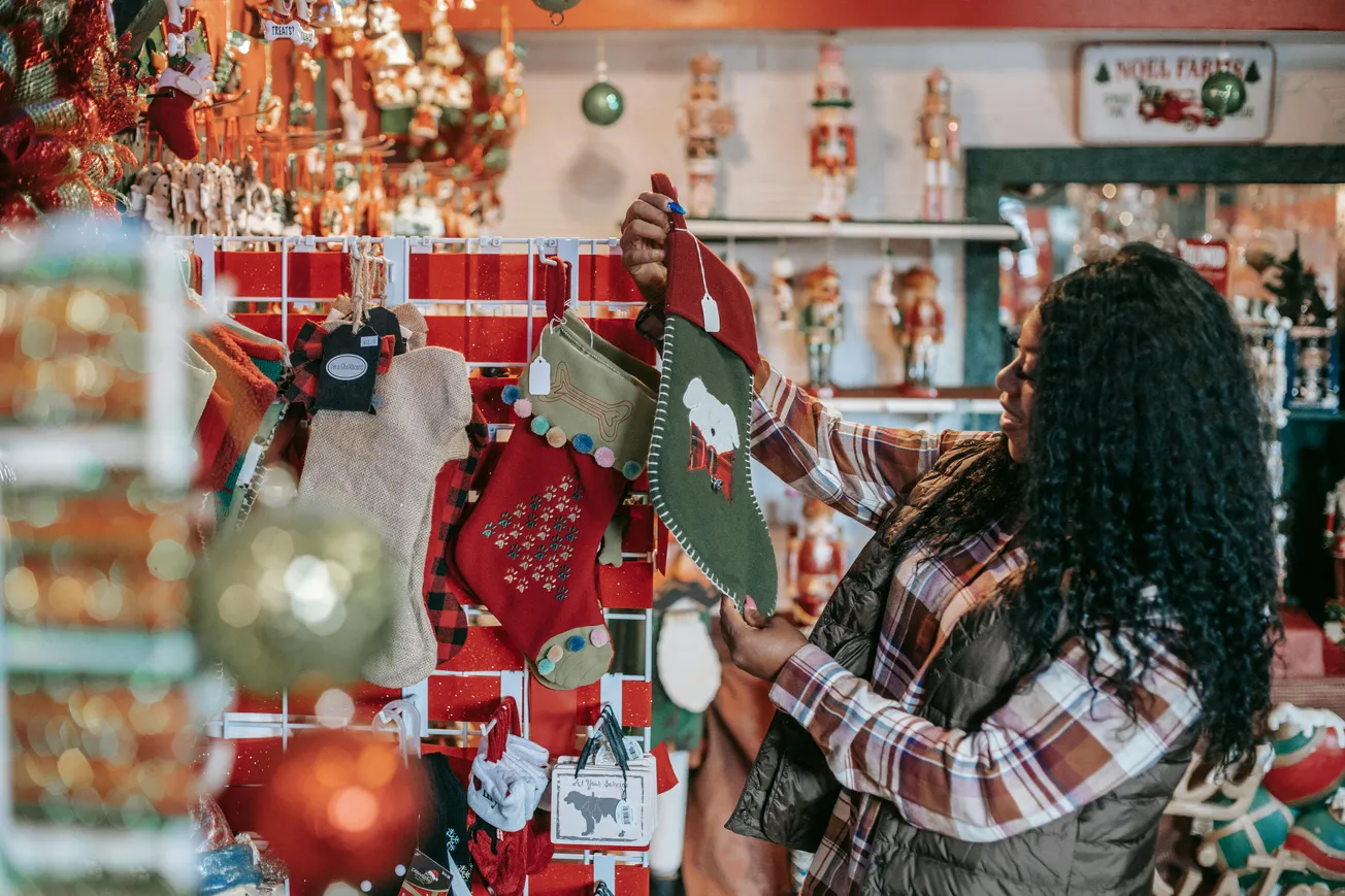 A woman with curly hair, in a plaid shirt, examines a green Christmas stocking in a festive store filled with holiday decorations and ornaments.