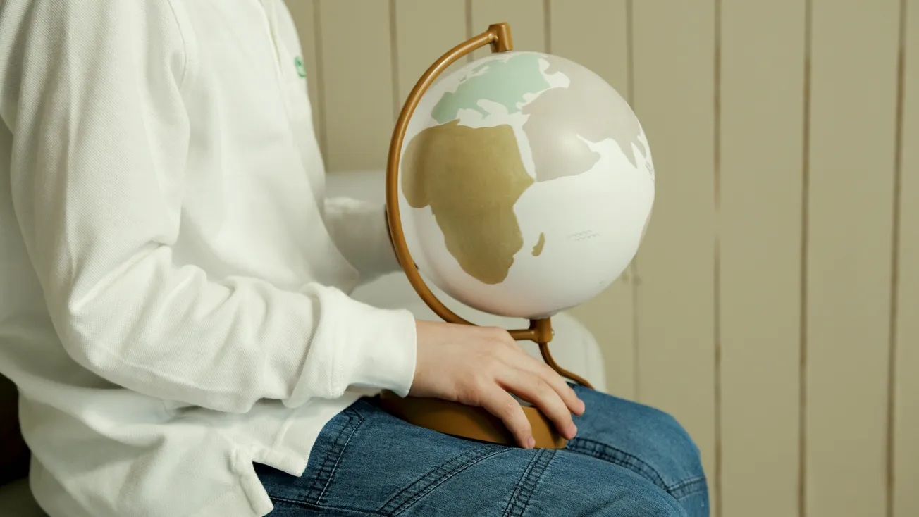 A person in a white shirt and jeans holds a small globe, focusing on Africa. The scene conveys curiosity and exploration, set against a wooden background.
