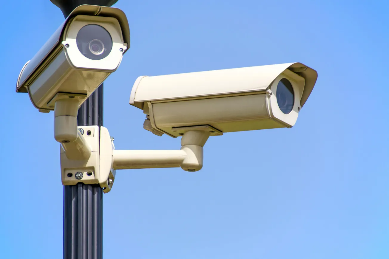 Two beige security cameras mounted on a black pole against a clear blue sky, indicating surveillance in a public area.