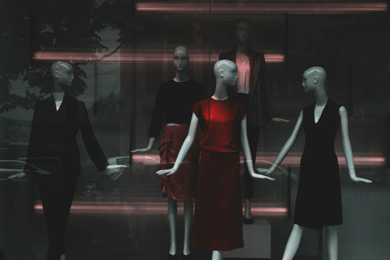 Five mannequins display outfits in a dimly lit store window. The central figure wears red, surrounded by outfits in darker tones, creating a stylish, moody ambiance.