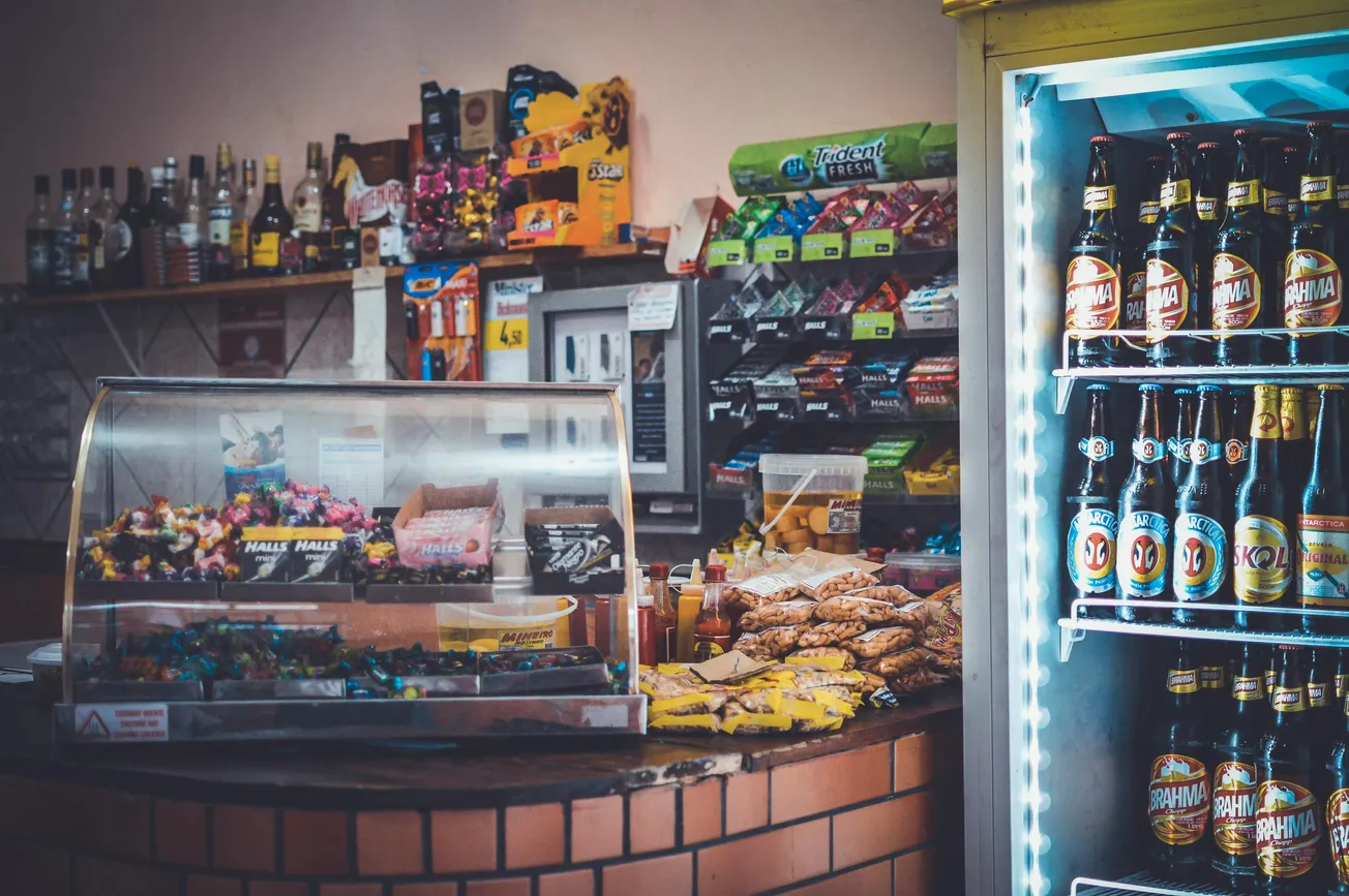 A cozy convenience store interior showcasing snacks, candy, and drinks. A fridge with bottled beverages and a counter with pastry and candy packs evoke a bustling, inviting atmosphere.