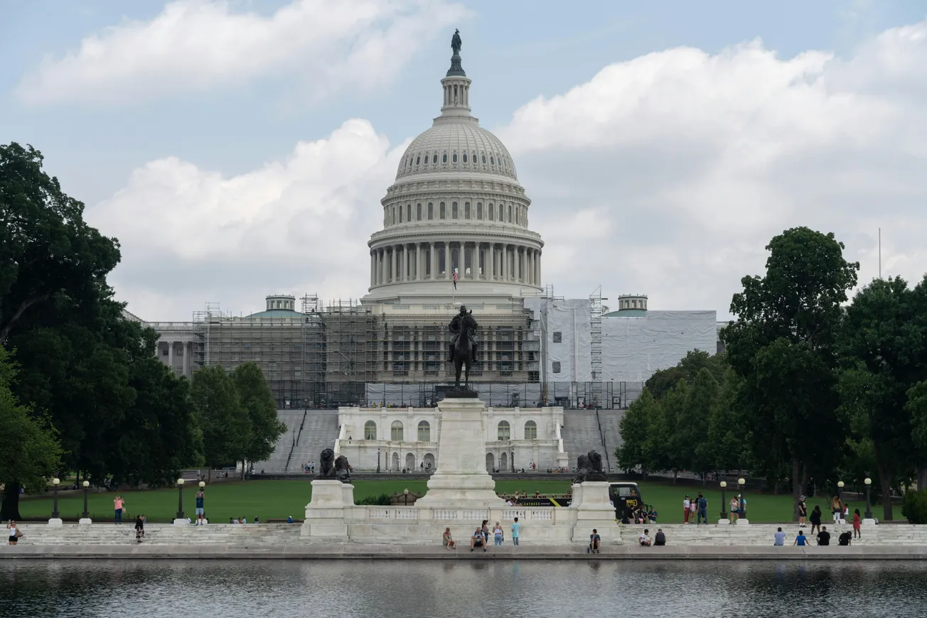 U.S. Capitol behind a statue and reflecting pool, framed by trees. Scaffolding covers parts of the building, with people and clouds adding lively elements.