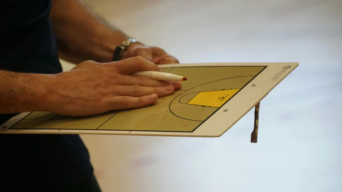 A person holds a basketball tactics board and pen, focusing on a play strategy, suggesting concentration and planning. The board is partially visible.