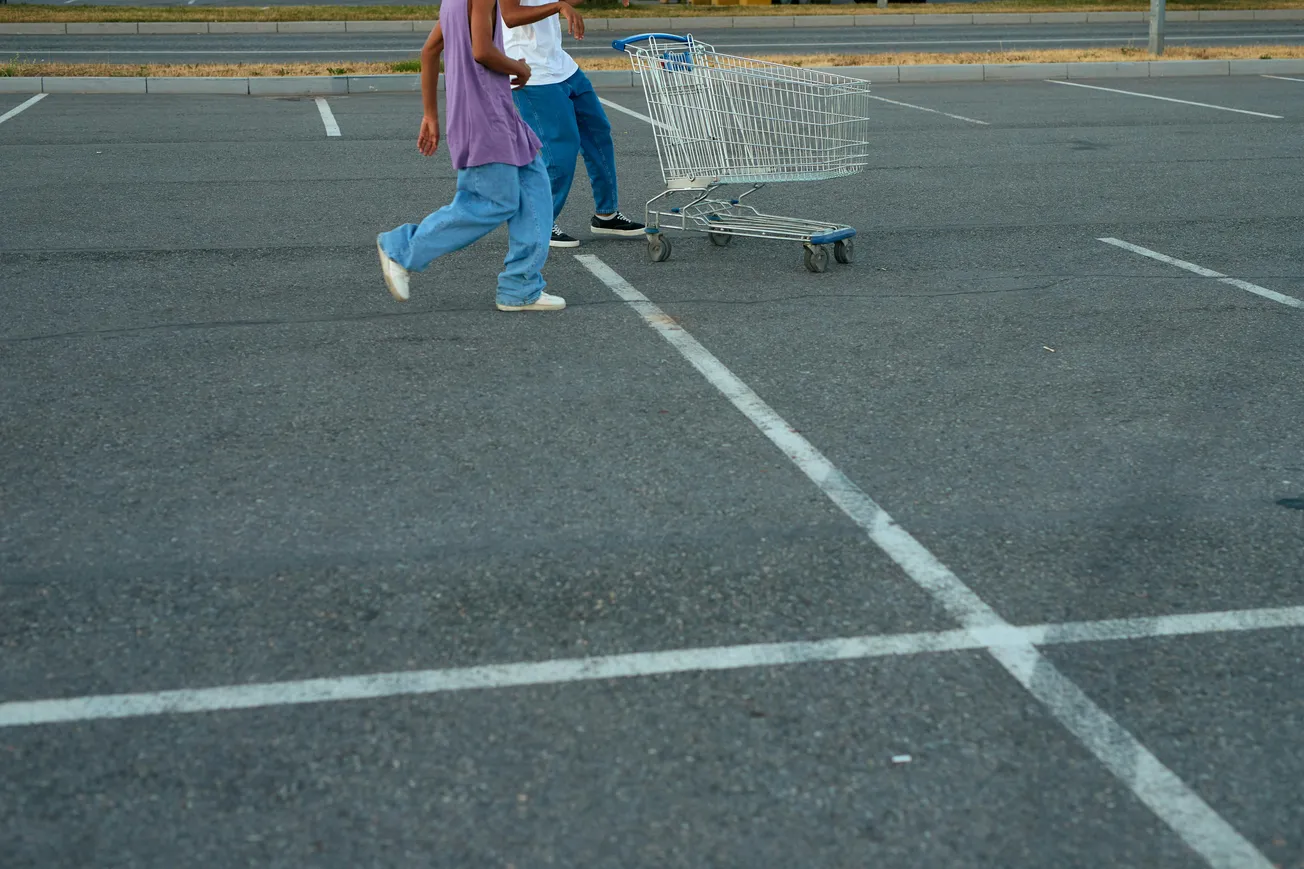 Two people walking briskly in an empty parking lot, pushing a shopping cart. They wear casual clothes, and the scene conveys a relaxed, everyday vibe.