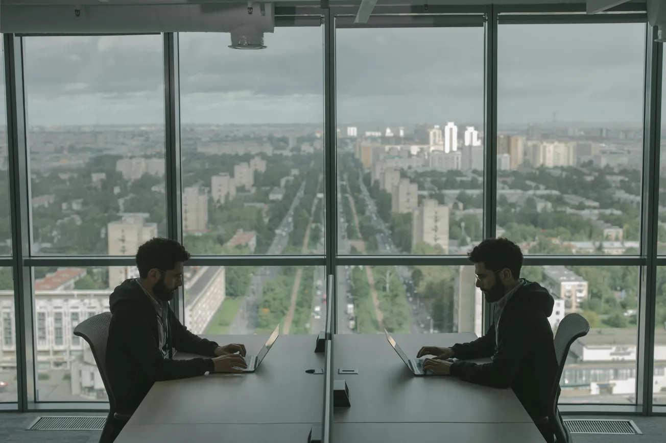 Two identical individuals sit at a table in a modern office, back-to-back, using laptops. A large window showcases a cityscape with a central road.