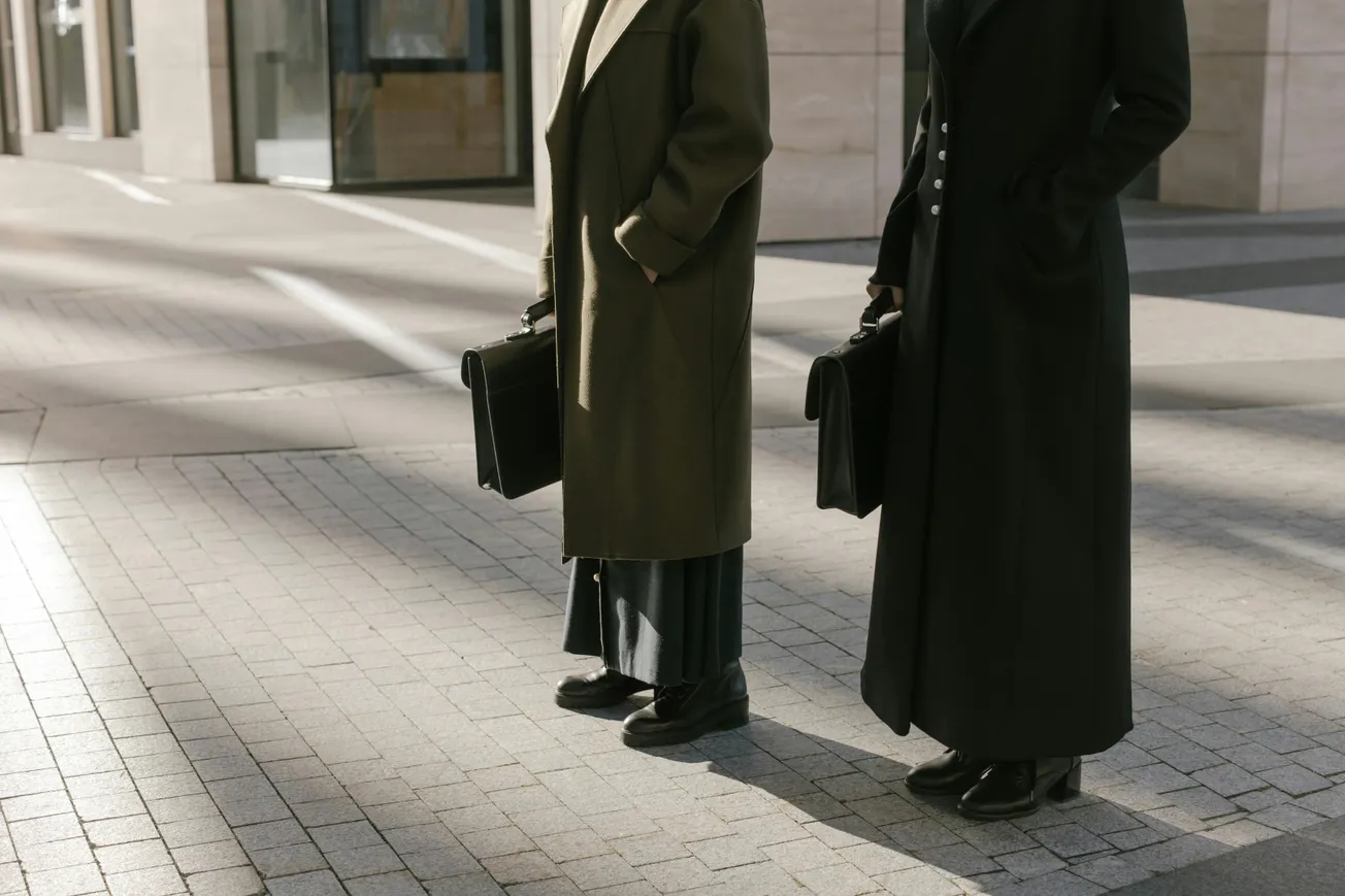 Two people in long coats stand on a sunlit, paved sidewalk holding briefcases. Their formal attire and posture suggest professionalism.