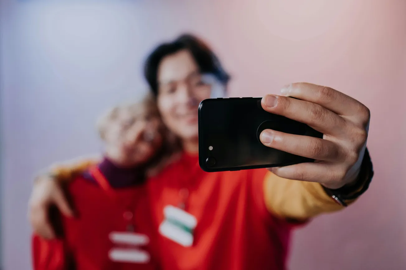 A smiling couple in matching red shirts takes a selfie. The photo is focused on the smartphone, with their faces slightly blurred in the background.