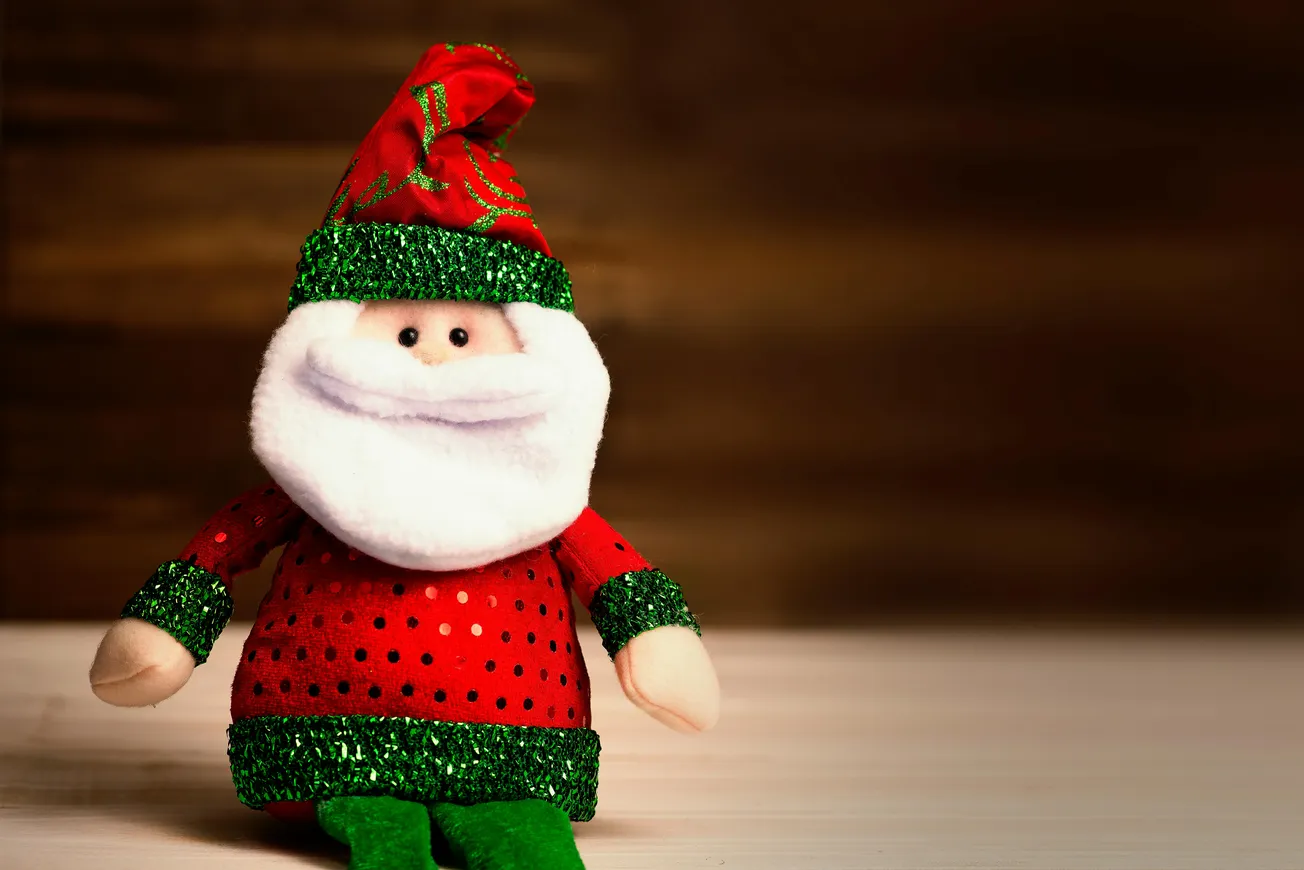 Plush Santa Claus toy in red and green outfit, sits on a light surface. It has a fluffy white beard and a joyful expression, against a warm, brown background.