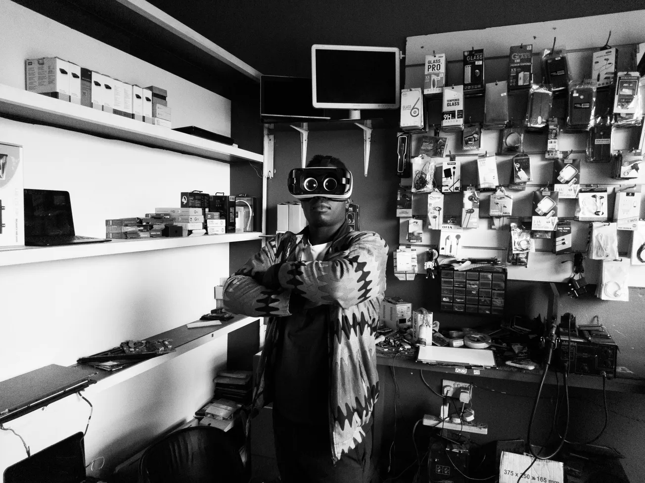 Person in VR goggles stands confidently with arms crossed in tech store. Shelves display boxed electronics and hanging accessories. Monochrome, futuristic.