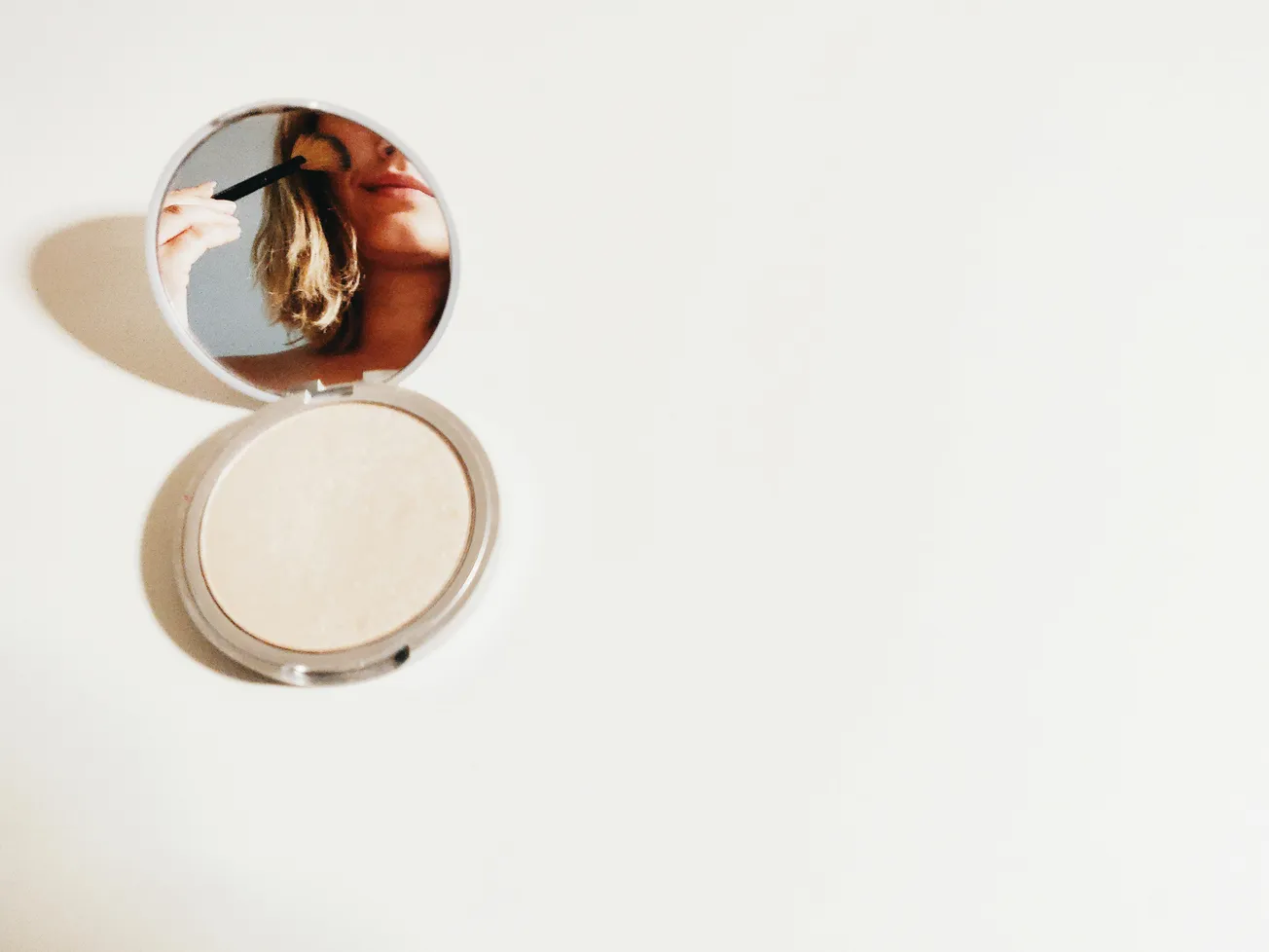Compact mirror on a white surface showing a reflection of a person applying makeup with a brush. The background is minimal and the mood is calm.