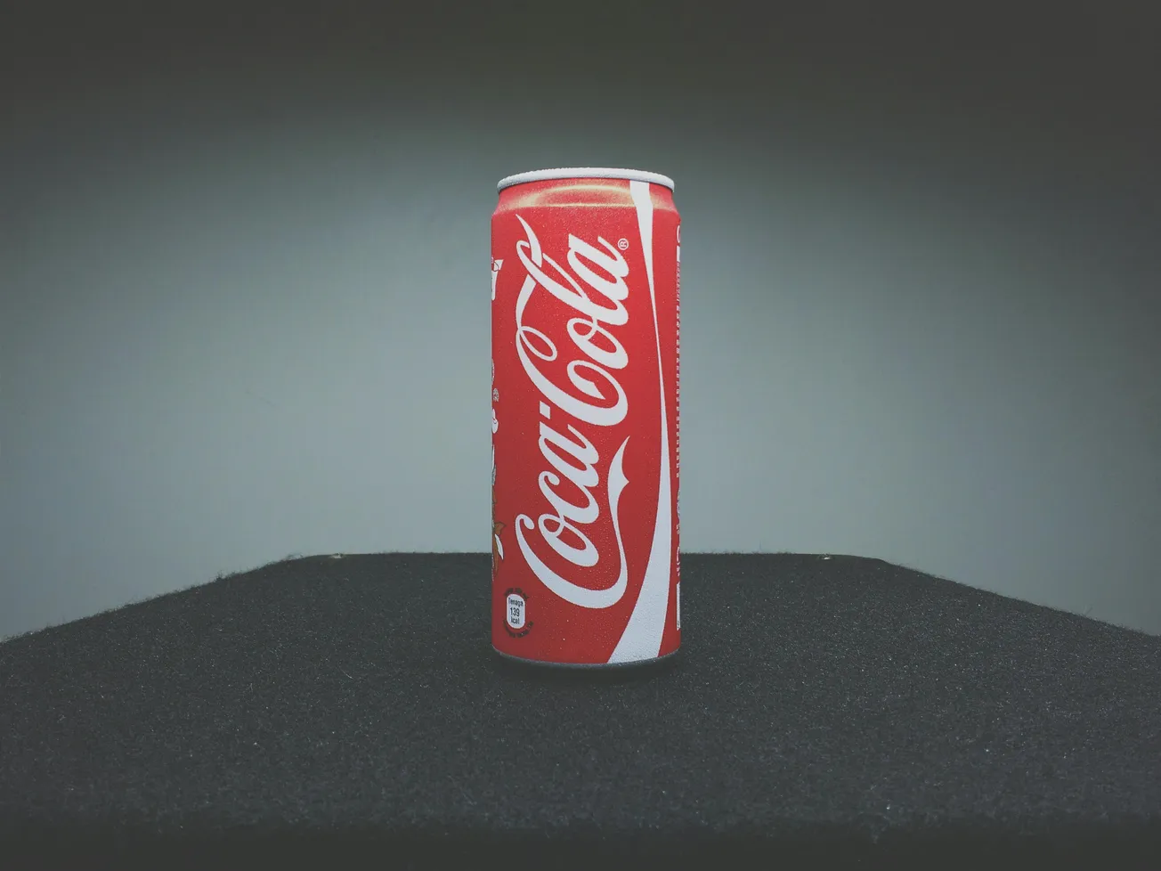 A red Coca-Cola can stands upright on a black surface. The can's white logo is prominent against the muted gray background, creating a classic, nostalgic feel.