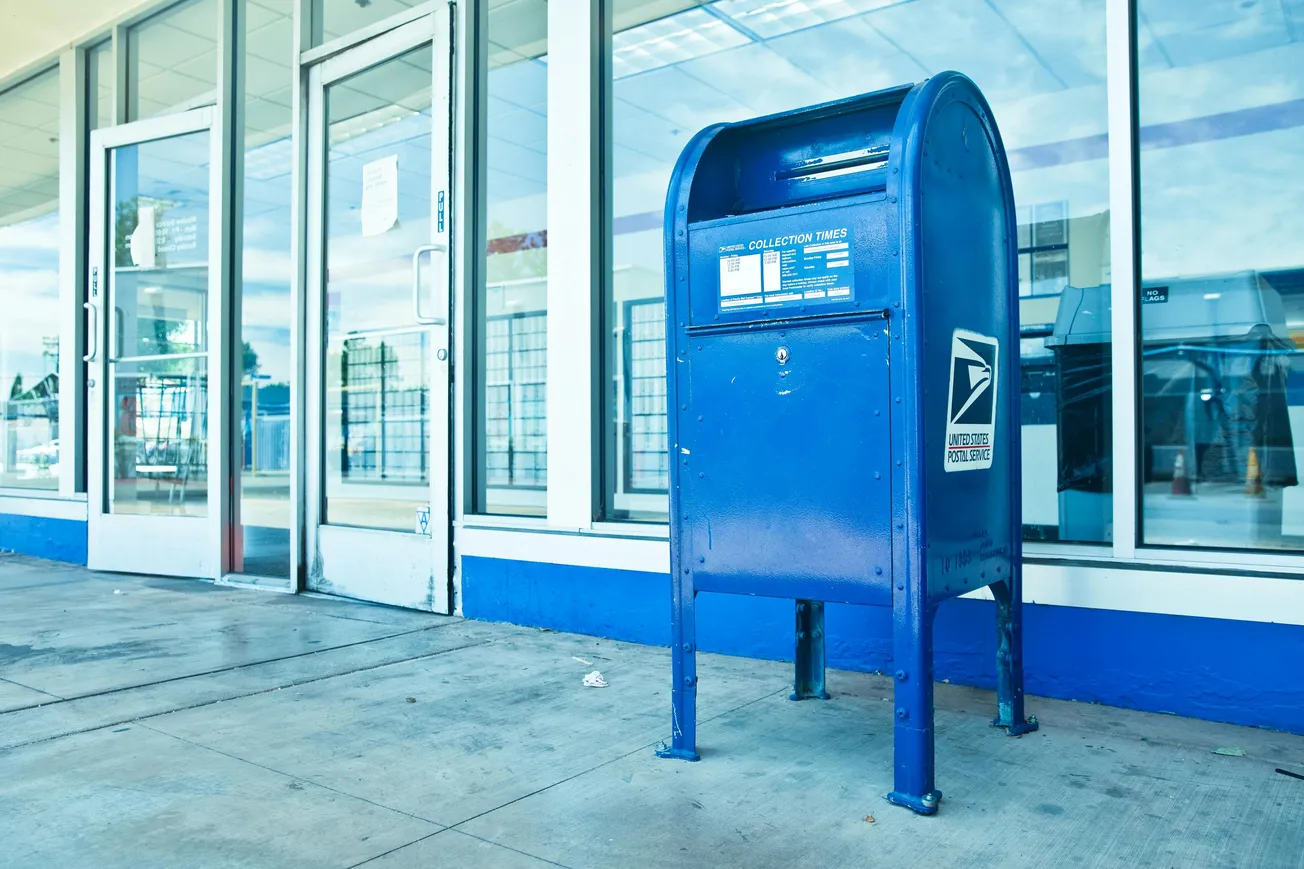 Bright blue USPS mailbox stands on a concrete sidewalk outside a glass-fronted building, evoking a sense of everyday urban life and accessibility.