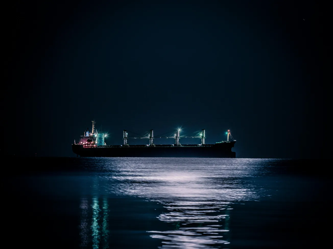 A cargo ship is silhouetted against the night sky, illuminated by soft lights. Its reflection shimmers across calm water, creating a serene, tranquil scene.