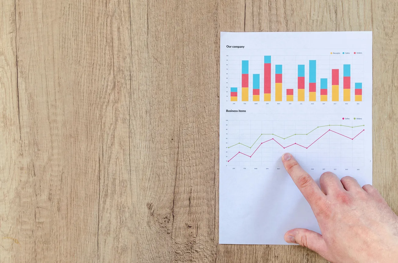 A hand points to a page with colorful bar and line graphs labeled "Our economy" and "Business trends" on a wooden table, conveying analysis and data evaluation.