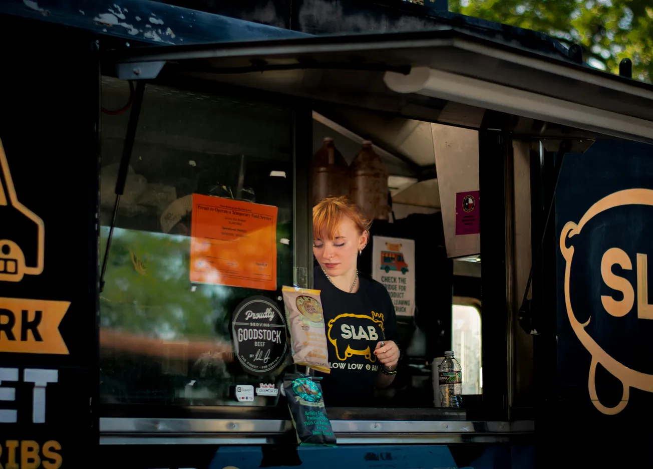 Young woman inside a food truck, reading a menu. She wears a "SLAB" shirt. The truck exterior has logos and colorful signs under sunny skies.