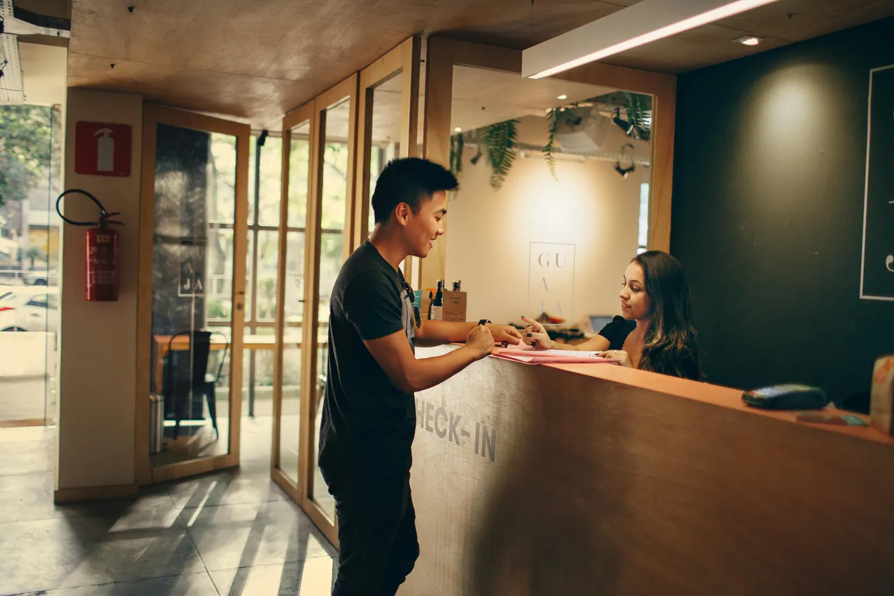 A man standing at a wooden reception desk is checking in with a receptionist. The room is warmly lit, creating a welcoming atmosphere.