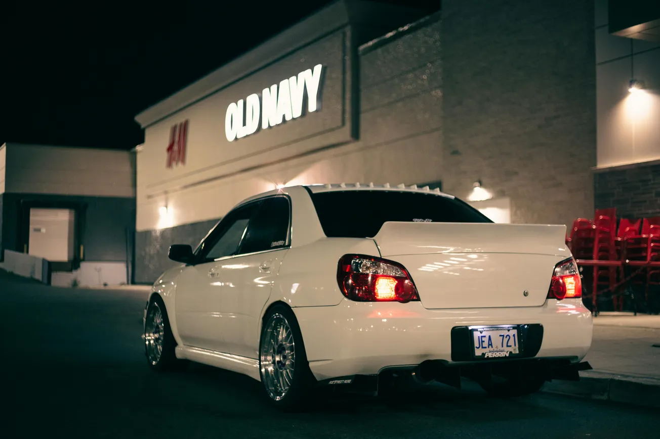 A white car is parked at night in front of an Old Navy store, with illuminated signs and stack of red chairs nearby, creating a serene urban scene.