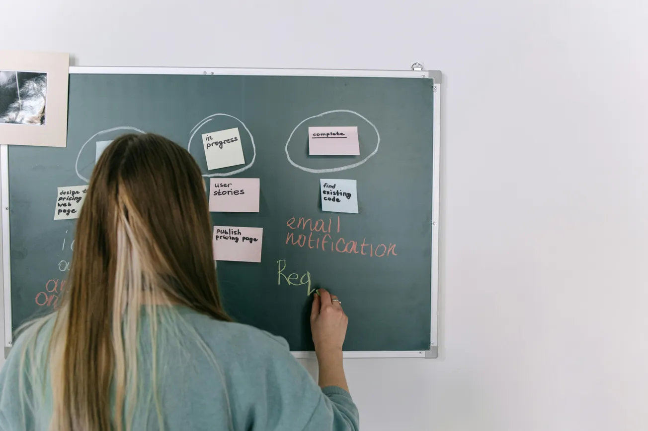 A woman with long hair writes on a chalkboard, planning projects with sticky notes and colored chalk. The scene conveys focus and organization.