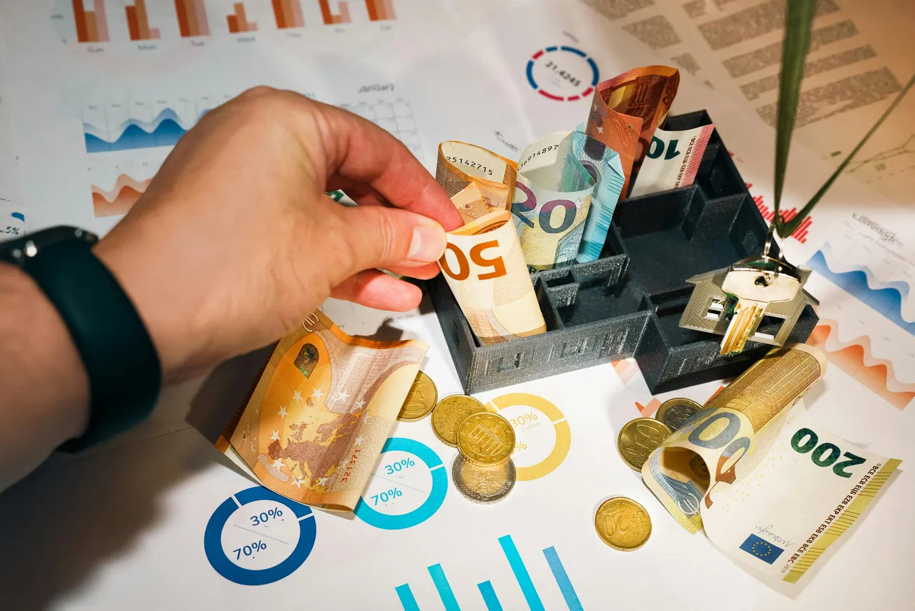 A hand arranges euro bills in a black cash tray surrounded by coins on financial charts, conveying themes of finance and money management.
