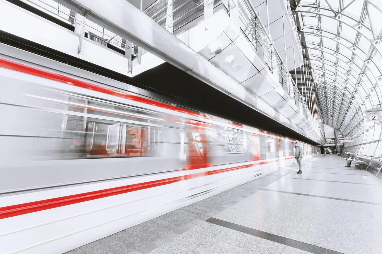 A sleek, modern train speeds past in a glass-arched station, creating a blurred motion effect. One person stands on the platform, adding a sense of anticipation.