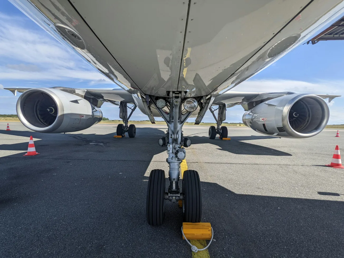 Underside view of a parked airplane showing landing gear and engines, with traffic cones on the tarmac under a clear blue sky.