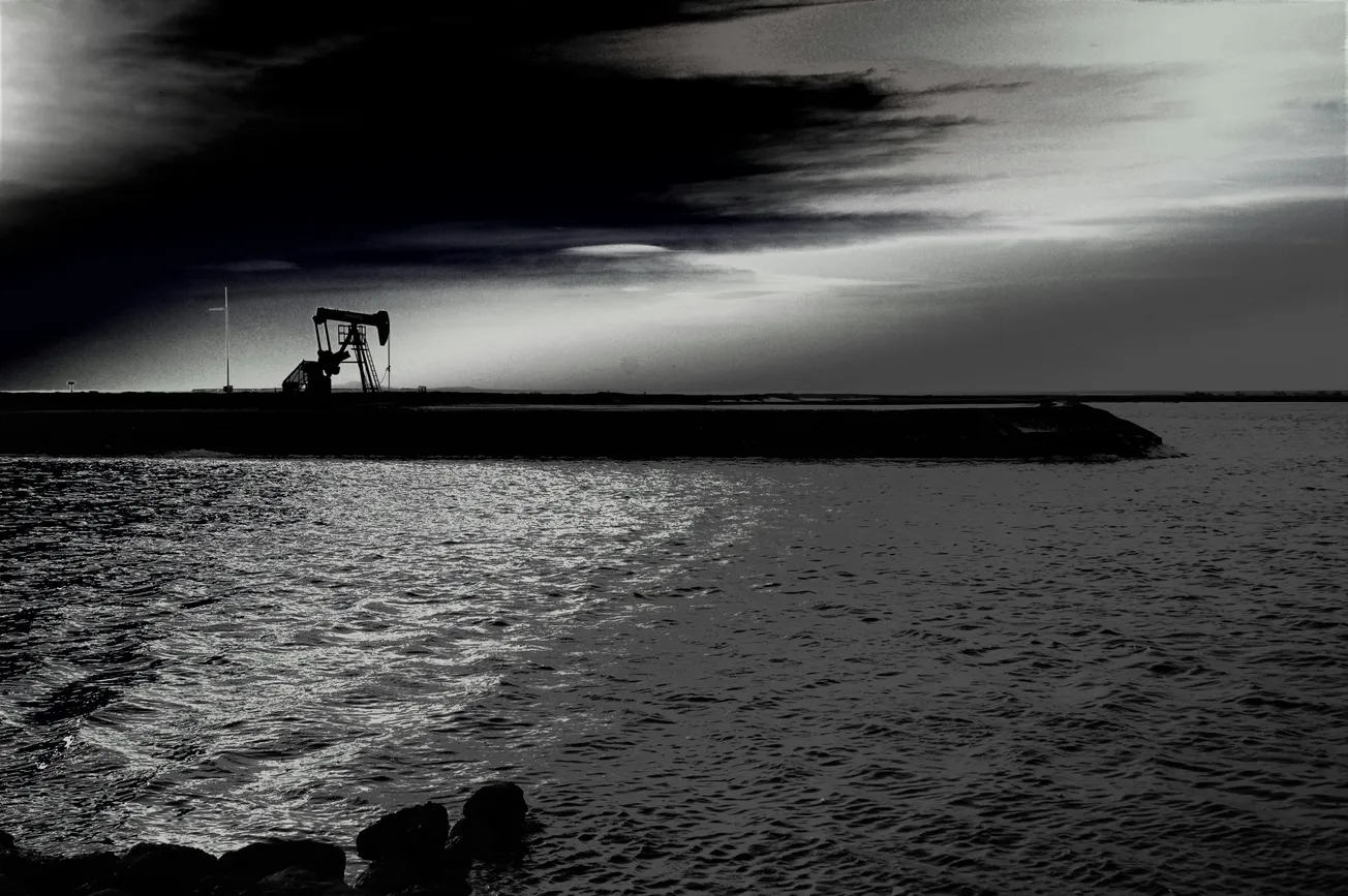 A lone oil pump silhouetted against a dramatic gray sky at dusk, near a reflective water body. The scene conveys solitude and industrial quietude.