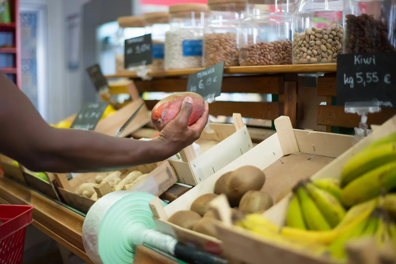 A person selecting a mango in a grocery store. The foreground shows bananas and kiwis, with jars of nuts in the background and price tags visible.