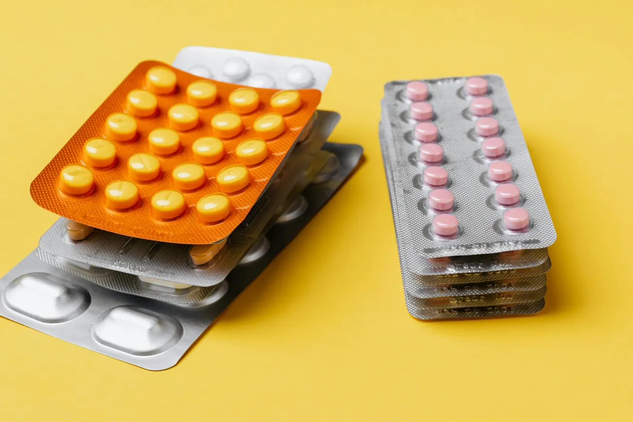 Blister packs of pills on a yellow background. One stack has orange packs with yellow pills; the other has silver packs with pink pills, conveying an organized, clinical feel.