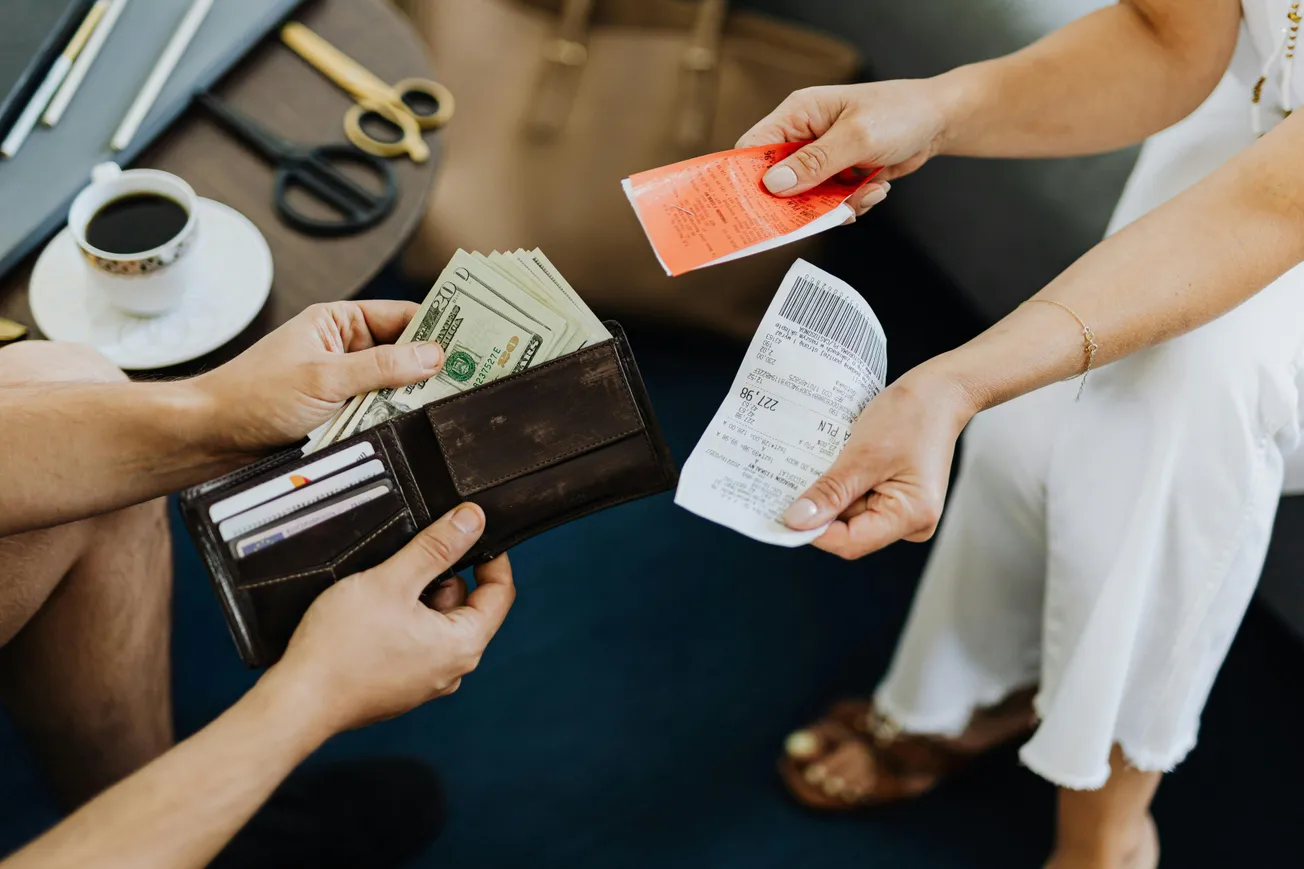 Two people exchange a brown wallet holding cash and receipts, seated indoors with a coffee cup on a nearby table, creating a business-like atmosphere.