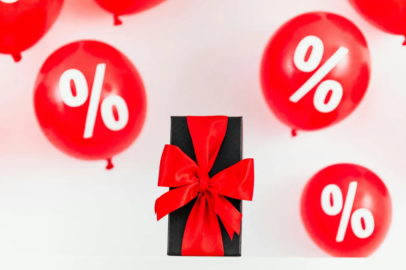 A black gift box with a bright red ribbon is centered among floating red balloons featuring white percentage symbols, conveying a festive sale or discount theme.