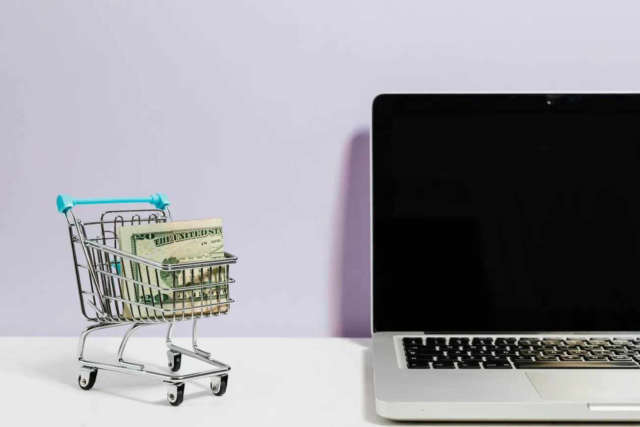 A small shopping cart filled with U.S. dollar bills is on the left beside a laptop with a blank screen on the right, set against a light purple background.