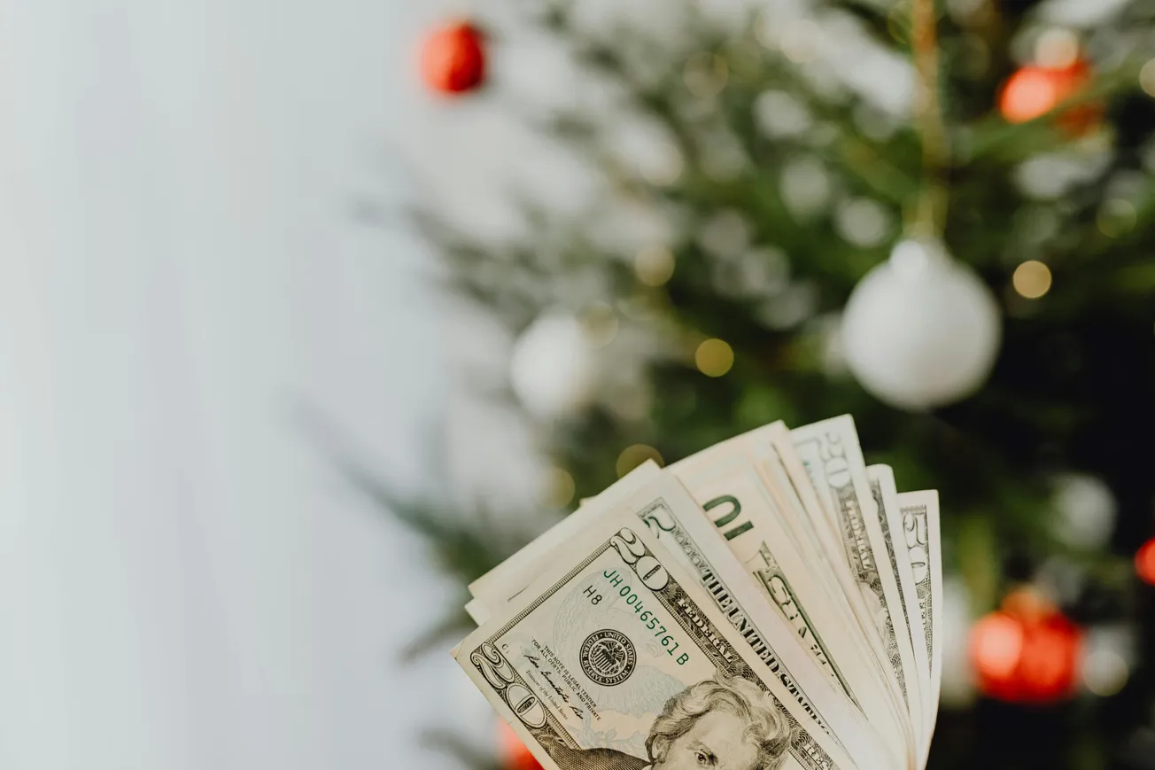 Close-up of fanned U.S. dollar bills held in front of a decorated Christmas tree with red and white ornaments, evoking a festive and prosperous mood.