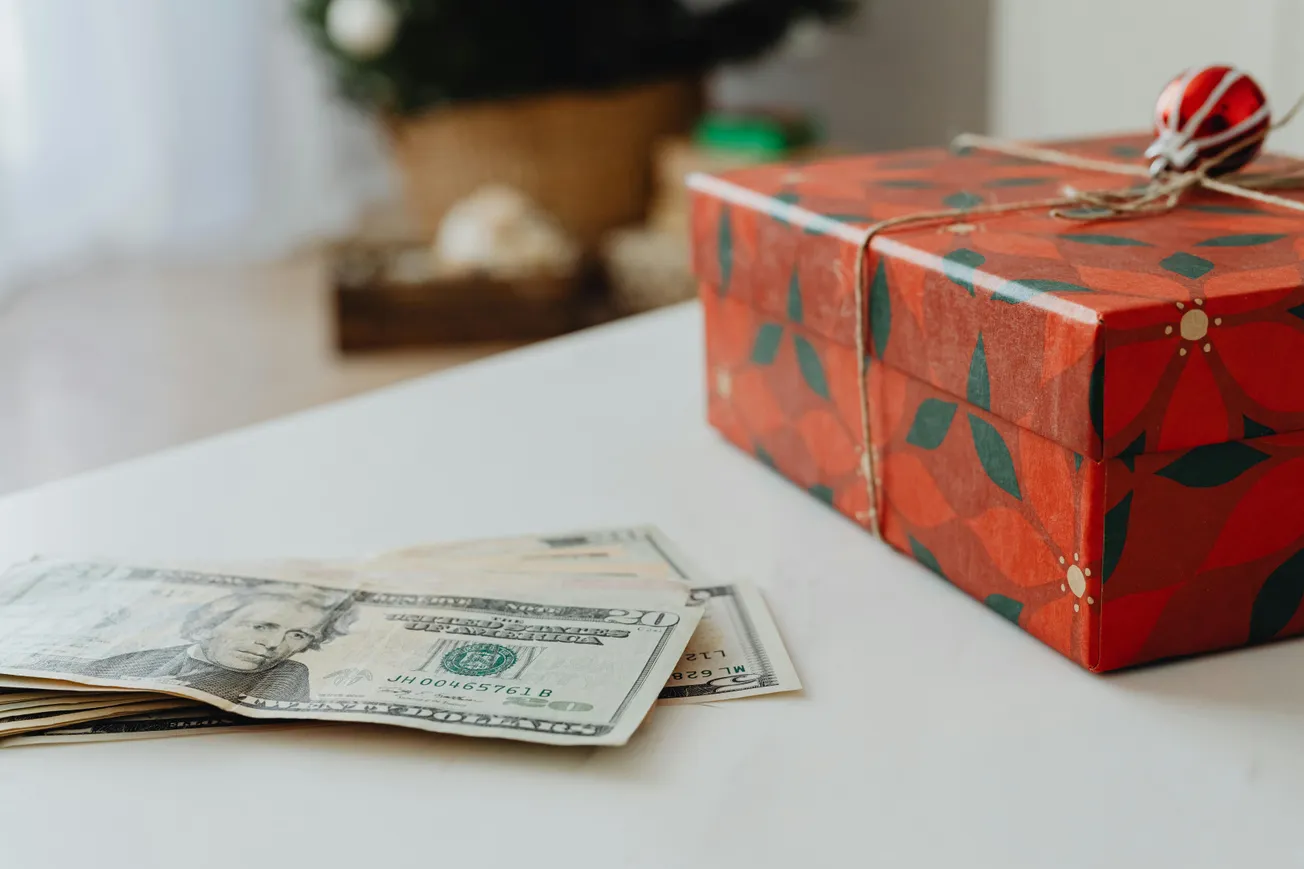 A festive red gift box with a floral pattern sits on a white surface next to a small stack of U.S. dollar bills, suggesting a holiday gift theme.