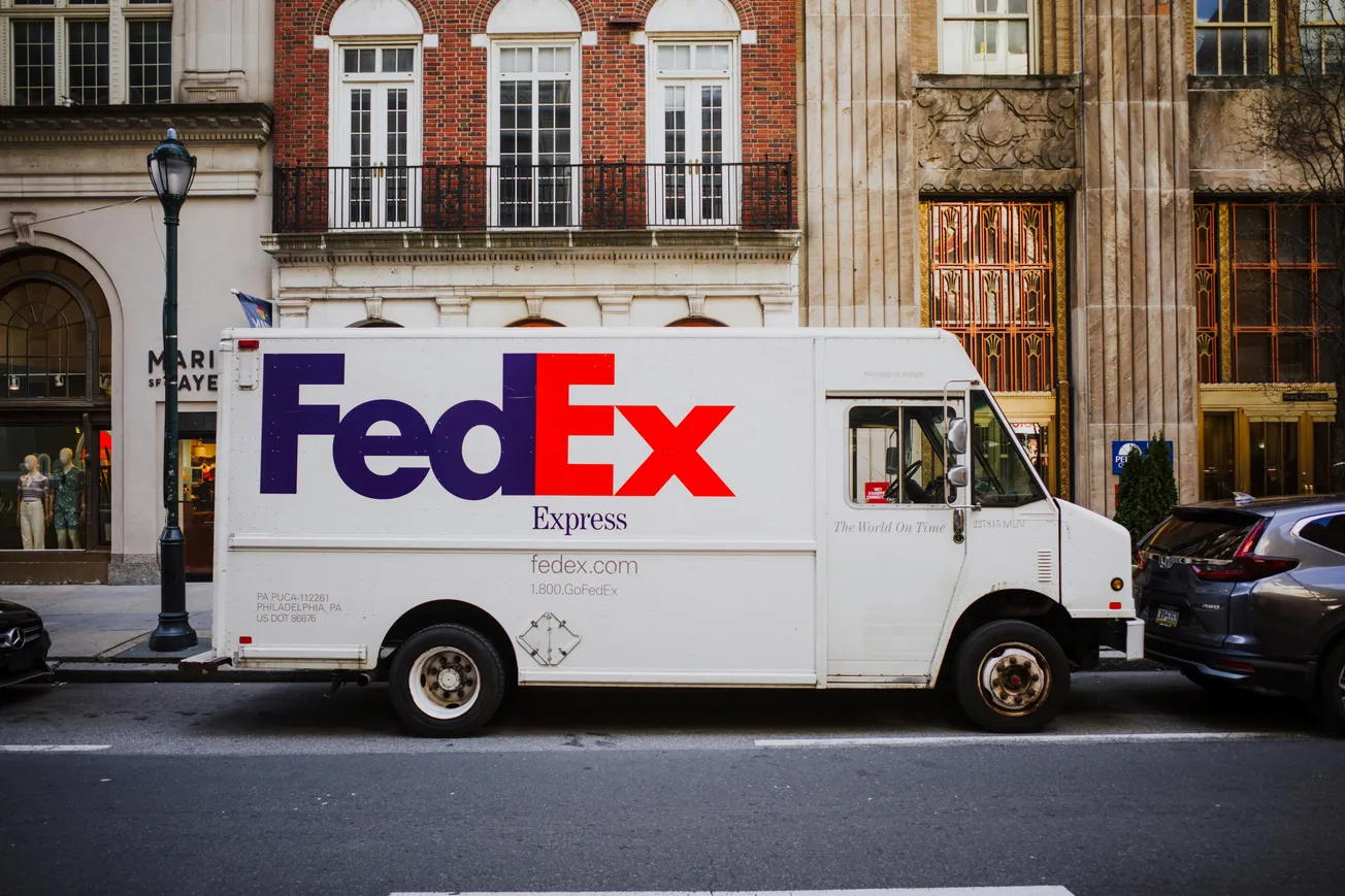 A white FedEx delivery truck is parked on a city street in front of an elegant brick and stone building with arched windows and decorative columns.