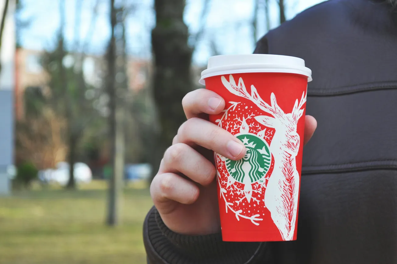 A hand holds a red Starbucks holiday cup with a white deer design in a park. The background shows blurred trees and grass, conveying a winter atmosphere.