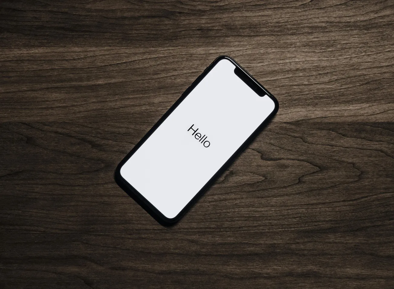 Smartphone on a wooden table, displaying "Hello" on its screen. The minimal design contrasts with the dark wood, creating a welcoming tone.