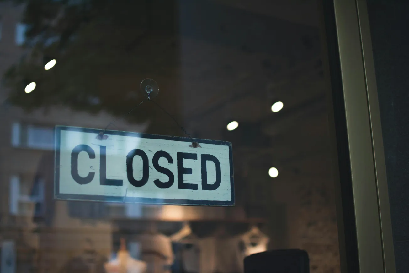 A "Closed" sign hangs on a shop glass door. The interior is dimly lit, and reflections show faint outlines of buildings, creating a calm, urban atmosphere.