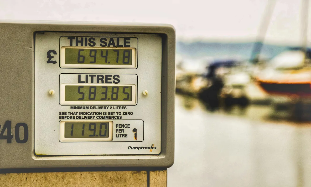 Close-up of a fuel pump display showing a sale amount of £694.78 for 583.85 liters at 119.9 pence per liter. Blurred boats in the background.