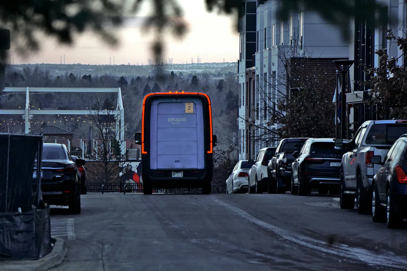 Autonomous Amazon delivery van with illuminated red edges drives down a quiet, car-lined street at dusk. Trees and hills are visible in the background.