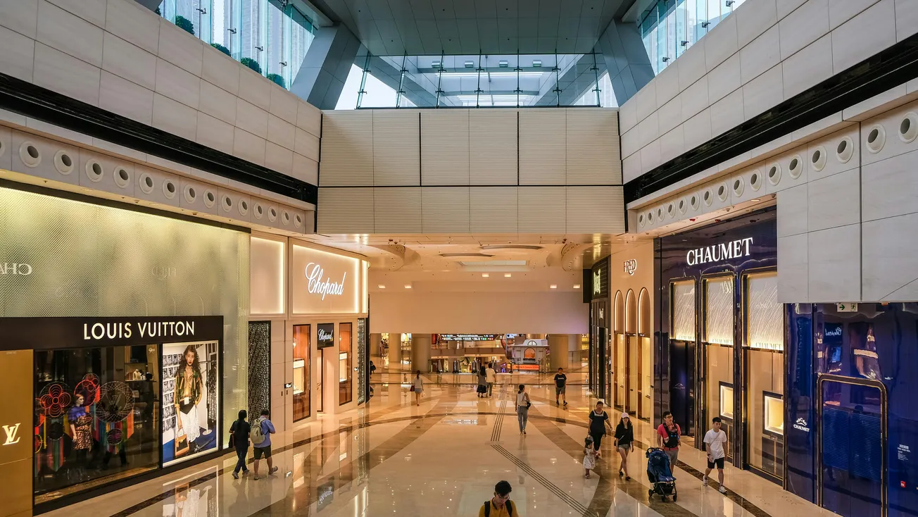 A modern, spacious shopping mall walkway with luxury stores like Louis Vuitton and Chaumet on both sides. Shoppers and bright, sleek design create an upscale vibe.