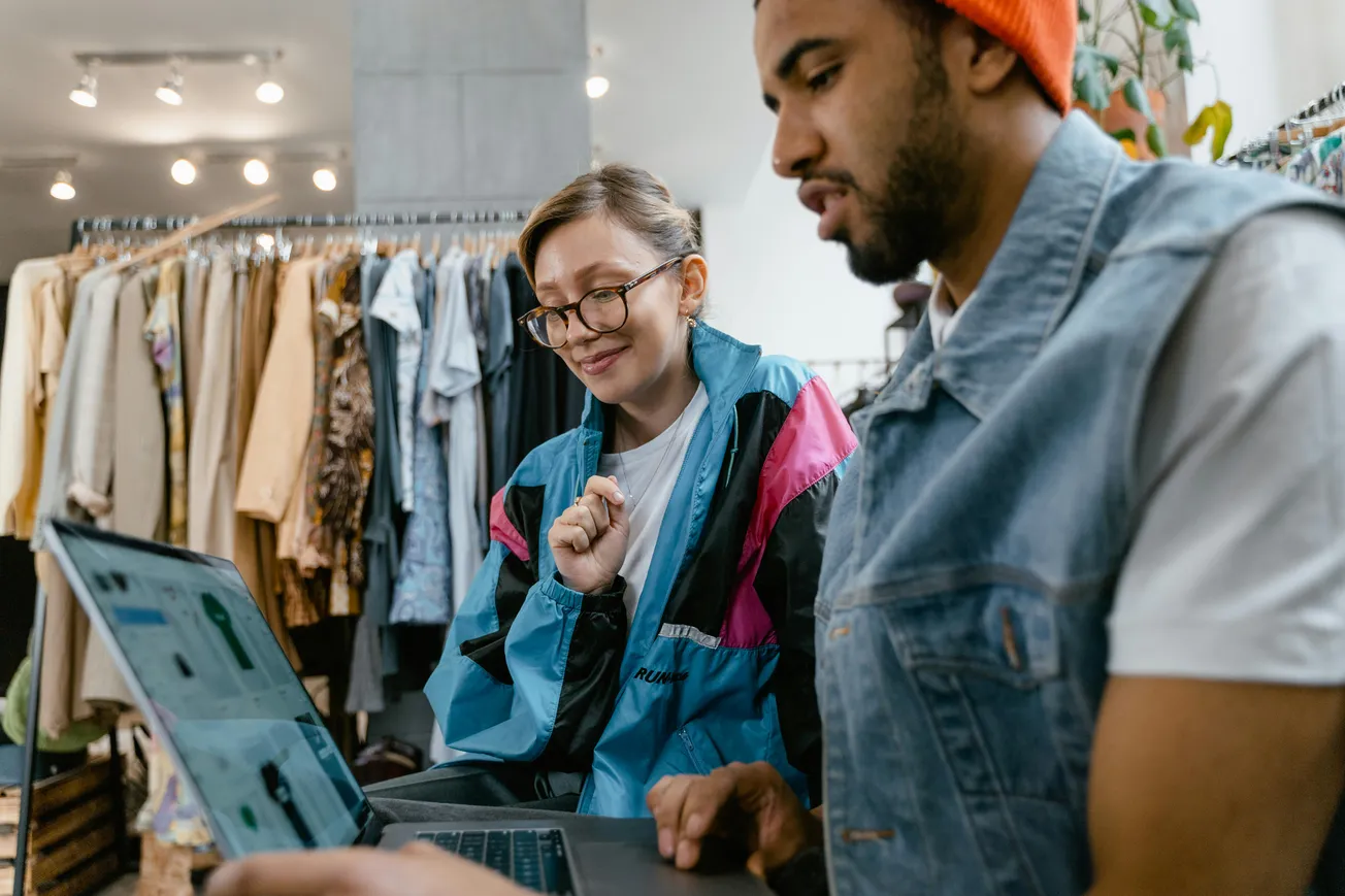 Two people in a clothing store, working on laptops. The woman wears glasses and a colorful jacket. The man, in a denim vest and beanie, types. Clothing racks are visible in the background.