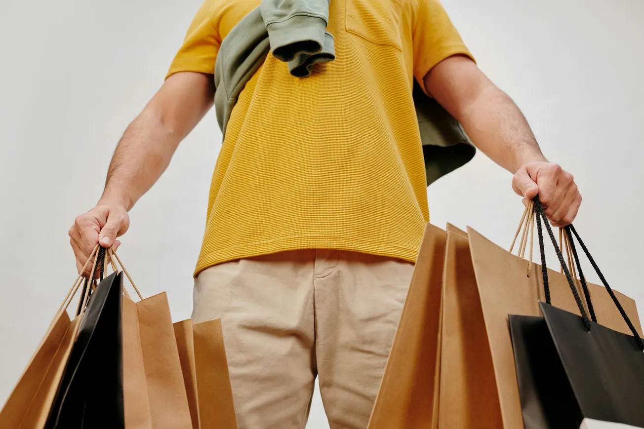Man in a yellow shirt holds multiple brown and black shopping bags. A jacket is draped over his shoulder, conveying a casual shopping vibe.
