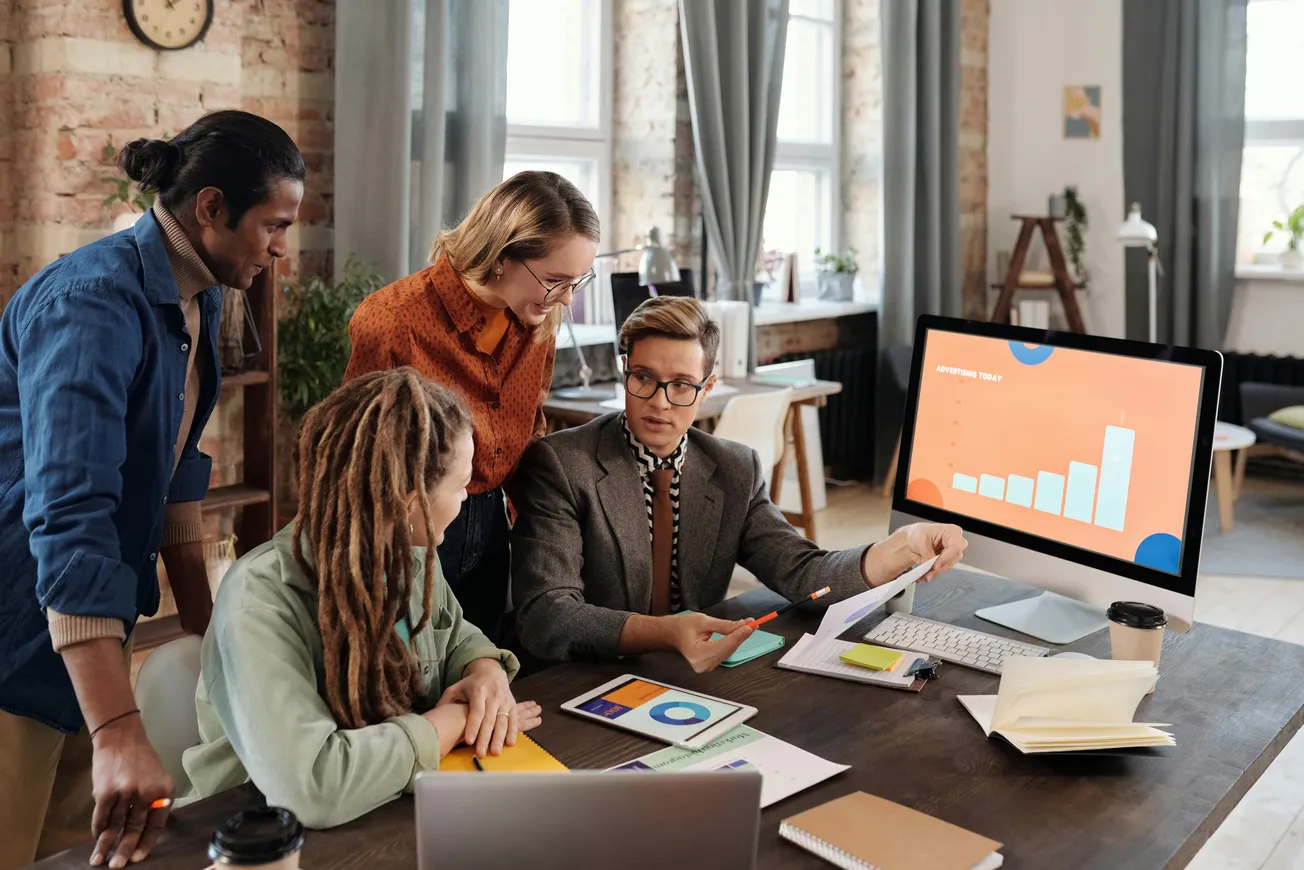 A diverse group of four people engaged in a collaborative meeting around a table with charts. A computer screen shows a bar graph, suggesting data analysis.