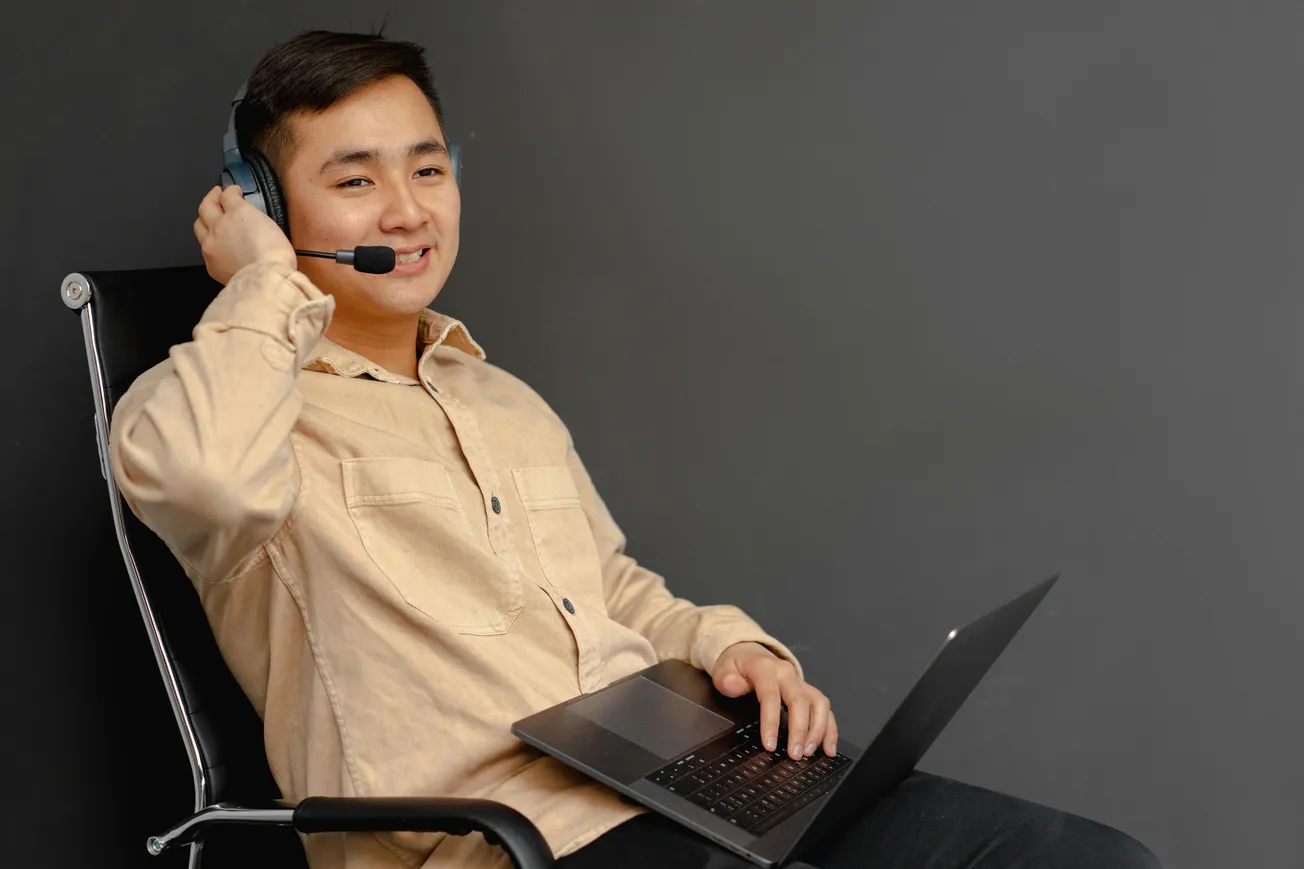 A smiling person wearing a beige shirt sits in a chair with a laptop, using a headset. The background is a plain dark wall, conveying a relaxed work-from-home vibe.
