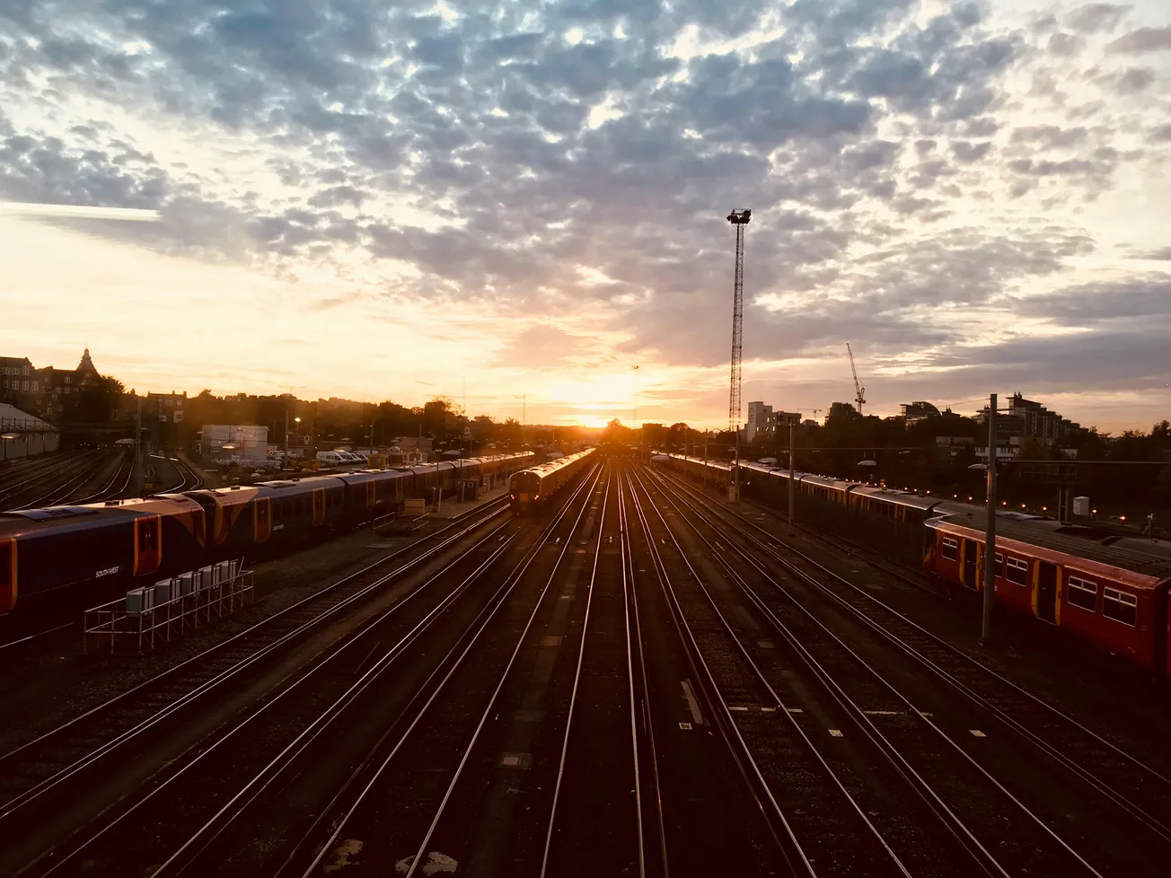 Sunset over railway tracks with trains on either side. The sky is filled with scattered clouds, casting a warm, peaceful glow across the scene.