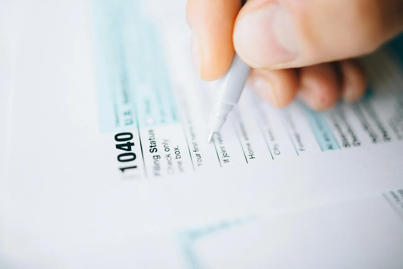 A close-up of a hand holding a pen, filling out a U.S. 1040 tax form. The scene conveys focus and organization, emphasizing tax season tasks.